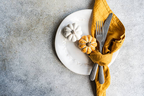 Top view of white ceramic plate with decorative pumpkins with silver fork and knife, wrapped in a mustard-colored napkin, placed on a textured gray concrete surface - ADSF48998