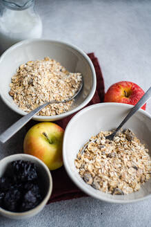 High angle of crop bowls with uncooked oatmeal placed on gray table near apples, milk jar and raisins - ADSF48999