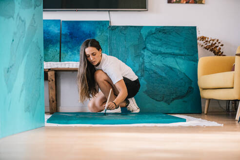 Young concentrated female artist in casual outfit in studio near blue painting and drawing on floor with paintbrush and watercolor paint - ADSF49002