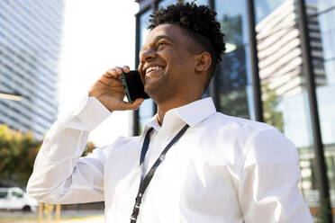 Low angle portrait of cheerful African American male entrepreneur in formal wear speaking on smartphone while standing on street and looking away - ADSF49025