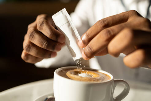 Crop closeup of anonymous black remote worker with hands adding sachet sugar to mug of hot tasty aromatic coffee against dark background in room - ADSF49028