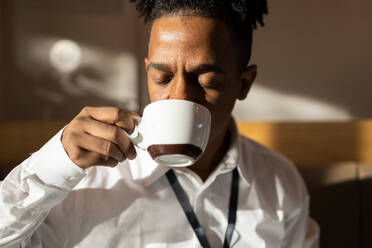 Portrait of pensive African American man drinking hot coffee with eyes closed while placed against bright blurred background of cafe - ADSF49029