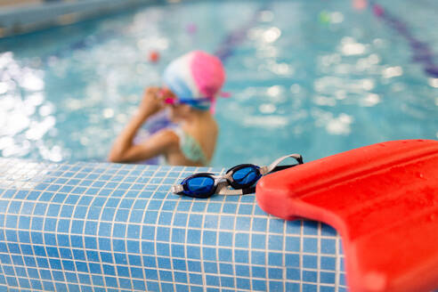 A child swimmer adjusts her blue goggles beside the pool, with a focus on a red kickboard and goggles in the foreground - ADSF49032