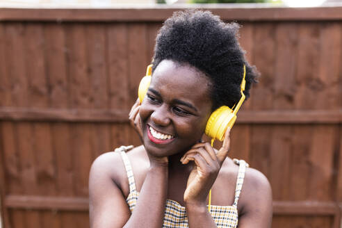 Lächelnde Frau, die vor einer Holzwand über Kopfhörer Musik hört - WPEF07830