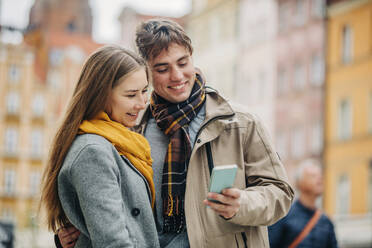 Smiling man sharing smart phone with girlfriend at street - VSNF01442