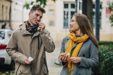 Smiling couple eating burgers standing at street - VSNF01448