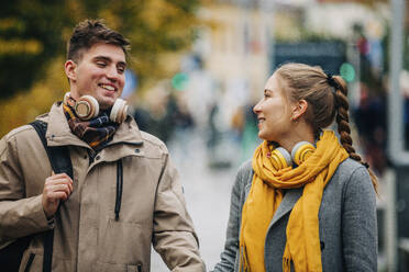 Glückliches Paar mit drahtlosen Kopfhörern, das gemeinsam auf der Straße spazieren geht - VSNF01450
