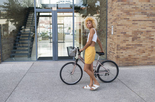 Young transgender woman standing with bicycle on footpath in front of building - VRAF00241