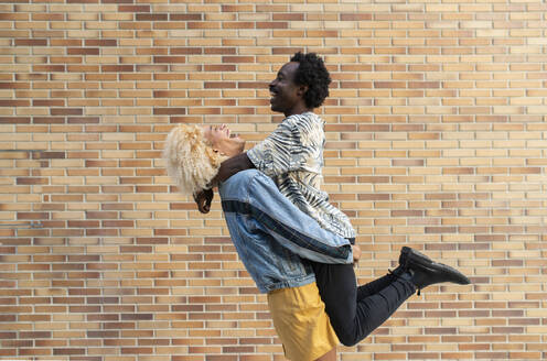 Cheerful transgender woman lifting boyfriend in front of brick wall - VRAF00249