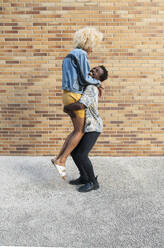 Boyfriend lifting transgender woman standing in front of brick wall - VRAF00251