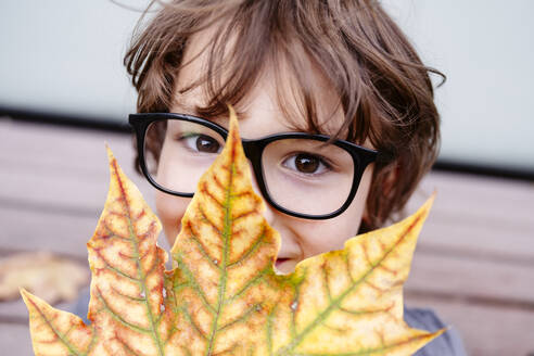 Junge mit Brille hält sich ein Blatt vor das Gesicht - EYAF02862