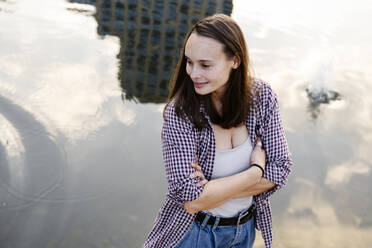 Smiling woman with arms crossed standing near lake - EYAF02866