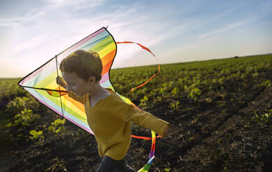 Happy boy holding kite and running in agricultural field under sky - MBLF00162