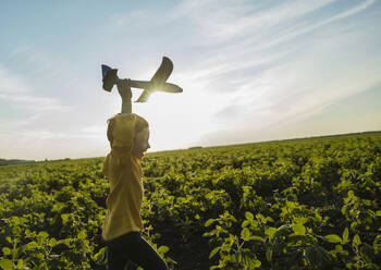 Fröhlicher Junge läuft mit Spielzeugflugzeug in landwirtschaftlichem Feld unter Himmel - MBLF00166