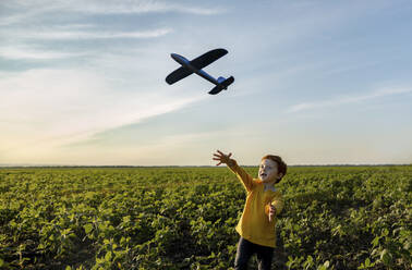 Carefree boy playing with toy airplane in agricultural field under sky - MBLF00167
