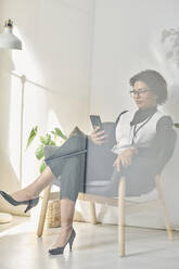 Businesswoman sitting in chair and using mobile phone seen through glass - DSHF01107