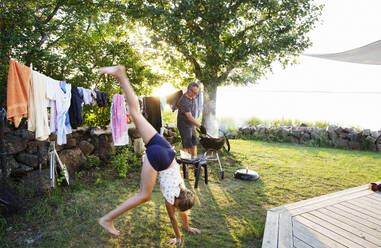 Girl doing cartwheel while her father cooks on barbecue - FOLF12392