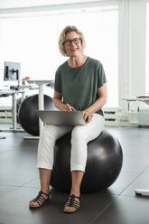 Woman using laptop on exercise ball in office - FOLF12535