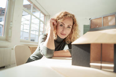 Smiling architect sitting with model house at desk - JOSEF22034