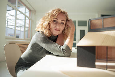 Smiling blond architect examining model house at desk - JOSEF22035