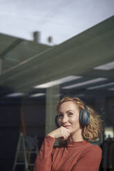 Smiling woman wearing wireless headphones seen through glass - JOSEF22063
