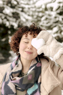 Smiling woman covering eye with heart shaped snow in winter - VIVF01253