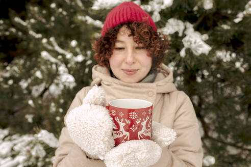 Lächelnde Frau mit Handschuhen und einer Kaffeetasse in einem Winterwald - VIVF01255