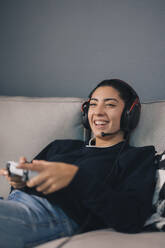 Cheerful young female in casual clothes and headset sitting on comfortable sofa while playing video game with joystick at home - ADSF49075