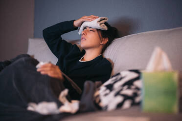 Sick young female in warm clothes with eyes closed while lying on cozy sofa with towel on forehead holding tissue paper and relaxing at home - ADSF49077