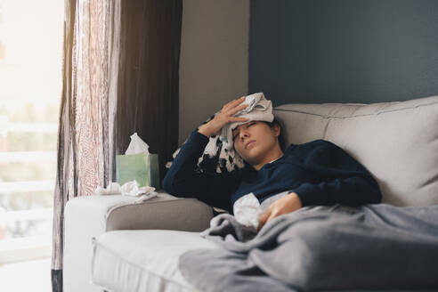 Sick young female in warm clothes with eyes closed while lying on cozy sofa with towel on forehead holding tissue paper and relaxing at home - ADSF49078