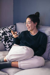 Smiling young female with hair bun looking at screen of mobile phone while relaxing on cozy sofa in daylight and reading text messages in living room - ADSF49079