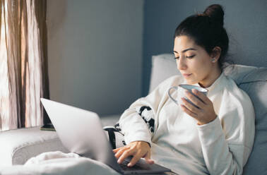 Side view of crop young female looking at screen of laptop while sitting on sofa and holding cup of hot coffee in living room at home in daylight - ADSF49084