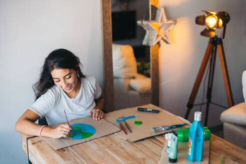 A cheerful young woman paints an earth design on cardboard, surrounded by painting supplies in a cozy room, highlighting environmental consciousness. - ADSF49087
