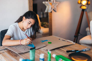 A cheerful young woman paints an earth design on cardboard, surrounded by painting supplies in a cozy room, highlighting environmental consciousness. - ADSF49089