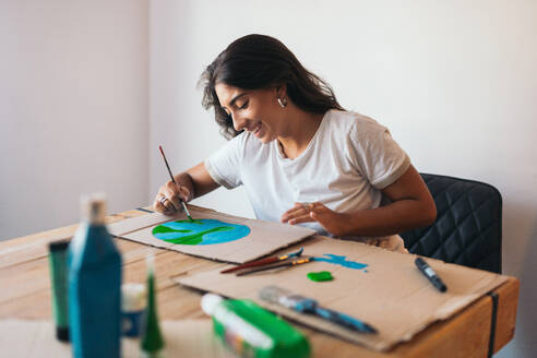 A cheerful young woman paints an earth design on cardboard, surrounded by painting supplies in a cozy room, highlighting environmental consciousness. - ADSF49090