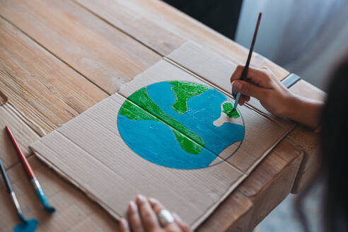 Close-up of a young woman's hands as she paints an earth design on cardboard, symbolizing environmental awareness and care. - ADSF49092