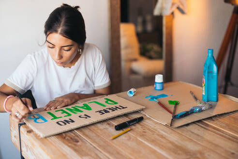 A cheerful young woman paints an text design on cardboard, surrounded by painting supplies in a cozy room, highlighting environmental consciousness. - ADSF49093