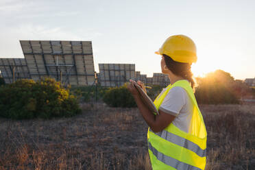 From behind a technical woman taking data next to some solar panels with a vest and protective helmet - ADSF49096