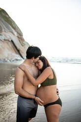 Side view of male kissing and touching tummy of pregnant woman in bikini leaning on his shoulder with closed eyes while standing against sea in summer evening - ADSF49107