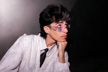 A young man with vibrant rainbow eyeshadow peering through clear glasses, set against a dark background, accentuated by a white shirt - ADSF49119