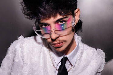 A young man with vibrant rainbow eyeshadow peering through clear glasses, set against a dark background, accentuated by a white shirt - ADSF49120