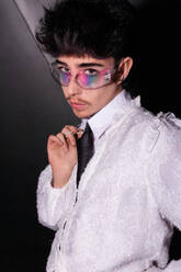 A young man with vibrant rainbow eyeshadow peering through clear glasses, set against a dark background, accentuated by a white shirt - ADSF49123