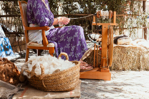 Unrecognizable woman in a purple dress sits by a spinning wheel, spinning yarn amidst raw wool and rural surroundings. - ADSF49129