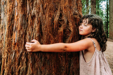 Seitenansicht eines süßen kleinen Mädchens, das sich tagsüber an einen alten Baumstamm in den Redwoods von Monte Cabezon schmiegt - ADSF49139