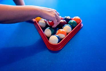 Crop anonymous person placing colorful pyramid of billiard balls in billiard triangle on blue pool table - ADSF49159