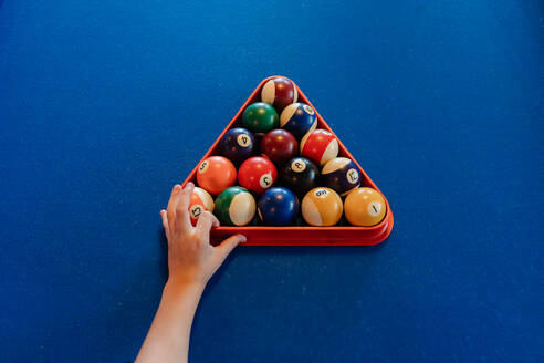 Top view of crop anonymous person placing colorful pyramid of billiard balls in billiard triangle on blue pool table - ADSF49160