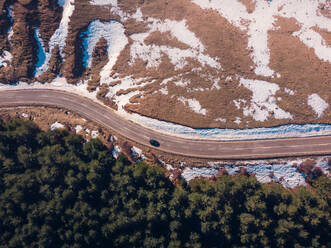 From above aerial view of a curving road surrounded by patches of snow and greenery, with a vehicle traveling along it in Cerler, Huesca, Spain - ADSF49169