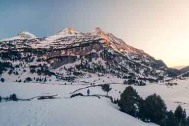 Rückenansicht einer nicht erkennbaren Person in einer blauen Jacke inmitten einer verschneiten Landschaft mit hohen schneebedeckten Bergen im Hintergrund und einem kleinen Dorf am Fuße der Berge. - ADSF49171
