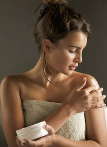 Young woman wrapped in a towel holding cream jar and rubbing her shoulder with body lotion - FSIF06670