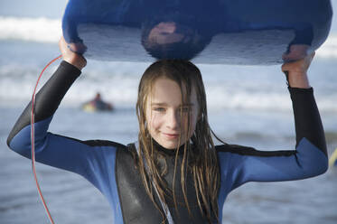 Lächelndes und nasses Mädchen, das ein Surfbrett auf dem Kopf trägt und aus dem Meer kommt - FSIF06679